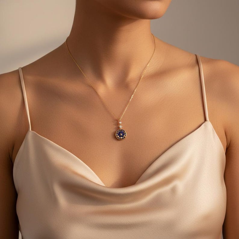 Puede incluir: Un collar dorado con un colgante circular. El colgante tiene una piedra central azul oscuro rodeada de piedras m&aacute;s peque&ntilde;as. El collar lo lleva una persona que lleva una blusa de sat&eacute;n.