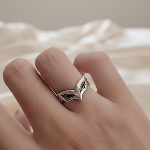 Op de afbeelding: Een zilverkleurige ring in de vorm van een masker, met donkere accenten. De ring wordt op een vinger gedragen, waarbij het ingewikkelde ontwerp wordt getoond. De maskerring is een uniek sieraad.