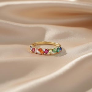 May include: A gold-colored ring with a row of colorful, rectangular gemstones. The gemstones are arranged in a rainbow pattern, with colors including red, orange, yellow, green, and blue. Small, clear stones are interspersed between the colored gems.