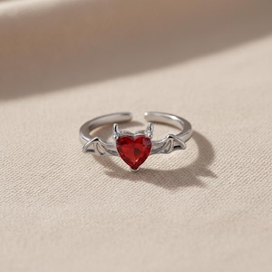 Może przedstawiać: Srebrny pierścionek z czerwoną, sercowatą kamieniem szlachetnym. Pierścionek ma diabelski motyw z małymi rogami i skrzydłami nietoperza. Pierścionek jest umieszczony na neutralnym tle.