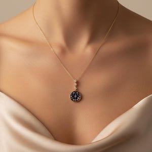 Puede incluir: Un collar dorado con un colgante con un dise&ntilde;o circular azul oscuro con peque&ntilde;os detalles blancos y azules. El collar tiene una cadena delicada y tres peque&ntilde;os detalles con forma de diamante encima del colgante.