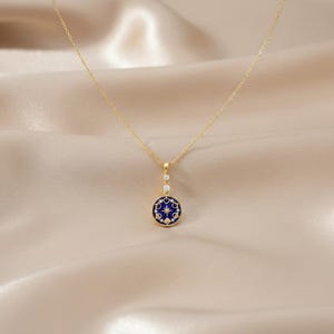 Puede incluir: Un collar dorado con un colgante circular. El colgante presenta un fondo de esmalte azul oscuro con un dise&ntilde;o de estrella y peque&ntilde;os detalles blancos. Dos peque&ntilde;as piedras transparentes est&aacute;n engastadas encima del colgante.