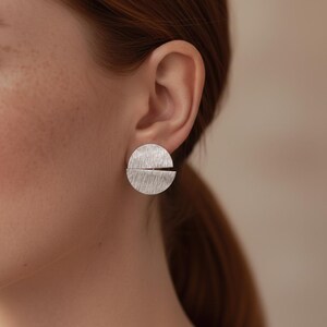 Peut inclure: Une paire de boucles d'oreilles argentées. Chaque boucle d'oreille est une demi-cercle texturé avec une fente horizontale. Les boucles d'oreilles sont rondes et ont une finition en métal brossé. Les boucles d'oreilles sont portées sur une oreille.