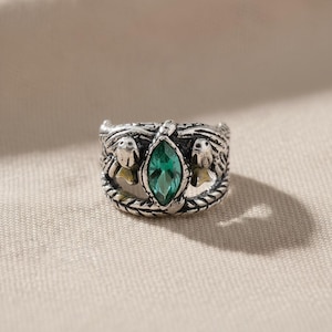 May include: A silver ring with a central emerald-cut green gemstone. The ring features detailed side designs of stylized figures and a braided band. The ring's design is intricate and ornate, suitable for a unique jewellery collection.