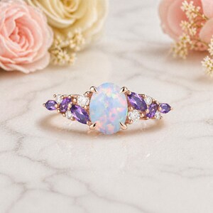 May include: A rose gold ring featuring a large oval opal center stone, surrounded by smaller purple and white gemstones. The ring is set against a marble surface, with soft pink and yellow flowers in the background.