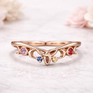 Könnte beinhalten: Roségoldener Ring mit keltischem Knotenmuster, besetzt mit rosa, blauen, gelben und roten Edelsteinen. Der Ring hat eine geschwungene Form und ist vor einem weichen, verschwommenen Hintergrund platziert.