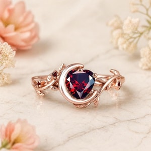 May include: Rose gold ring featuring a heart-shaped, deep red gemstone. The ring has a crescent moon design and delicate leaf accents. The ring is set on a marble surface, with soft pink and white flowers in the background.