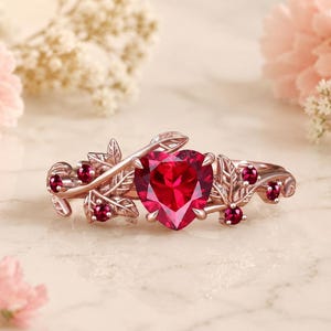 May include: A rose gold ring featuring a large heart-shaped red gemstone at the center, flanked by smaller round red gemstones and intricate metal leaf motifs along the band, creating a botanical design with a polished metallic finish.