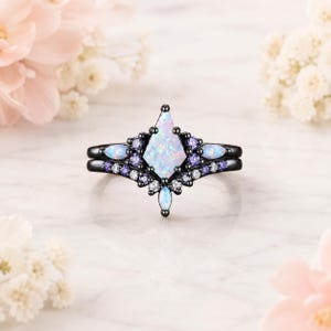 May include: A black metal ring set featuring a central kite-shaped opal, accented by smaller marquise-cut opals and round purple and clear stones. The design includes a curved band that frames the central stone with a symmetrical arrangement.