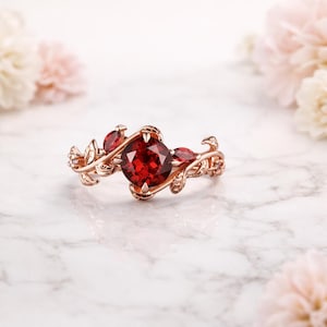 Puede incluir: Anillo de oro rosa con una gema roja oscura grande y redonda como punto focal, flanqueada por piedras rojas más pequeñas y delicados detalles en forma de hojas. El anillo se encuentra sobre un fondo suave y borroso con acentos florales.