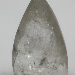 May include: A clear, triangular crystal with a pointed apex. The translucent crystal features internal textures and inclusions, set against a white backdrop. The smooth, polished form suggests a gemstone or decorative item, with a height of approximately 7 cm.