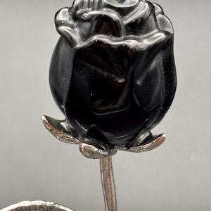 May include: A black rose sculpture with a polished, glossy finish. The rose is set on a silver-colored stem with detailed leaves. The sculpture is a decorative item, possibly for display.