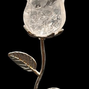 May include: A clear quartz rose sculpture with a silver stem and leaves. The rose is intricately carved, showing the natural crystalline structure of the quartz. The leaves have detailed veining. The sculpture is set against a black background.