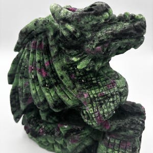 May include: A carved stone dragon head sculpture. The sculpture is green with red and black detailing. The dragon features detailed scales and a textured surface. The sculpture is set against a white background.