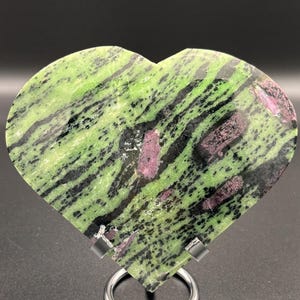 May include: A heart-shaped stone sculpture with a polished surface. The stone features a vibrant green base color with black and pink veining. The sculpture is displayed on a silver metal stand, showcasing the natural beauty of the stone.