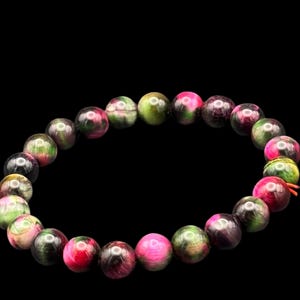 May include: A bracelet made of round beads in shades of pink, green, and black. The beads are strung together to form a flexible, circular shape. The bracelet is displayed against a black backdrop.