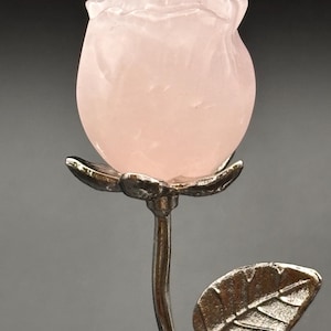 Op de afbeelding: Een roze roos sculptuur met een zilveren steel en blad. De roos is gesneden uit een doorschijnend, lichtroze materiaal, met gedetailleerde bloemblaadjes. De steel en het blad hebben een metallic zilveren afwerking, wat een contrast creëert.