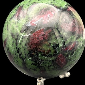Könnte beinhalten: Eine polierte Kugel aus grünem Zoisit mit Rubin-Einschlüssen, die auf einem dekorativen Ständer ruht. Der Edelstein weist ein gesprenkeltes Muster aus tiefroten und schwarzen Flecken auf einem leuchtend grünen Hintergrund auf. Die Kugel ist hochreflektierend.