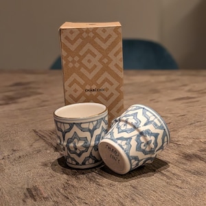 Puede incluir: Dos pequeñas tazas de cerámica con un patrón geométrico azul y blanco, una en posición vertical y otra inclinada. El borde de la taza tiene un ribete dorado. Una caja de cartón con un diseño geométrico está detrás de las tazas. El texto "CHABI CHIC" es visible.