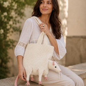 May include: A cream-colored crocheted tote bag shaped like an opossum. The bag features a textured body, four legs with pink feet, a long pink tail, a pointed snout with black eyes, and white whiskers. It includes two top handles for carrying.