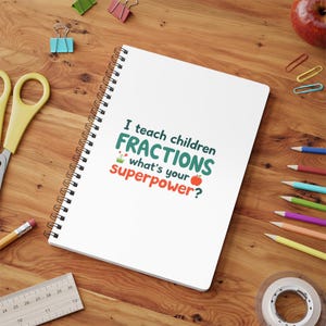 May include: A white spiral-bound notebook with the text "I teach children fractions, what's your superpower?" on a wooden desk. The notebook is surrounded by pencils, scissors, paper clips, a ruler, and an apple.