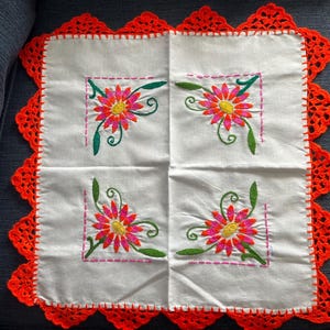 May include: White square cloth with four embroidered floral designs in each corner. The flowers are pink and yellow with green leaves and stems. The edges are trimmed with a bright orange crocheted border.