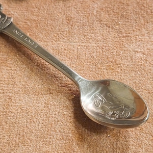 May include: A silver souvenir spoon with a decorative handle and bowl. The handle is adorned with a crest and the word "LUCERNE". The bowl features an embossed design. The spoon rests on a textured, brown surface.
