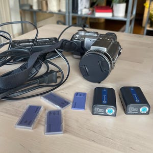 May include: A silver Sony digital camcorder with a black lens cap, a black power adapter with a cord, two rectangular black rechargeable batteries, and four small rectangular memory cards in clear plastic cases arranged on a light surface.