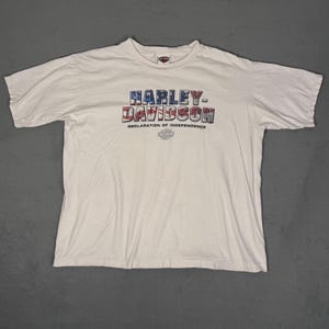 May include: Off-white Harley-Davidson t-shirt. The front features the Harley-Davidson logo in red, white, and blue, with "DECLARATION OF INDEPENDENCE" below. The shirt is made of a soft material and has short sleeves.