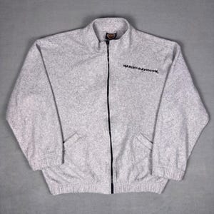 Könnte beinhalten: Eine hellgraue Fleecejacke mit Stehkragen und durchgehendem schwarzen Reißverschluss. Der Schriftzug Harley-Davidson ist in Schwarz auf der linken Brust gestickt. Die Jacke hat lange Ärmel mit elastischen Bündchen und einen elastischen Saum.