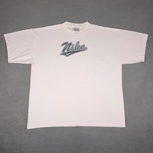 May include: White t-shirt with the Nike logo in a blue and gray design across the chest. The shirt has short sleeves and a crew neck. The shirt is laid flat on a gray surface.