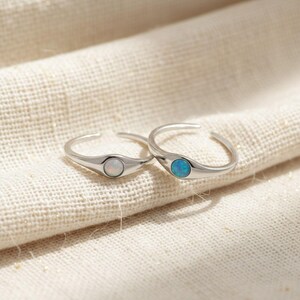 Puede incluir: Dos anillos de plata con diseño abierto. Un anillo presenta una piedra de ópalo blanco y el otro una piedra de ópalo azul. Los anillos se muestran sobre una tela texturizada de color blanquecino.