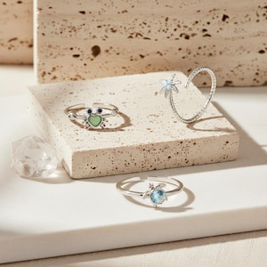 Könnte beinhalten: Eine Auswahl an Silberringen. Ein Ring hat ein Krabben-Design mit einem grünen herzförmigen Körper und blauen Augen. Ein weiterer Ring hat ein Palmen-Design, und der dritte hat einen blauen Edelstein.