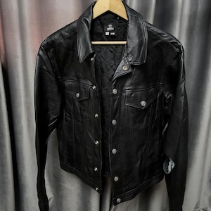 Puede incluir: Una chaqueta de cuero negra con cuello en punta, mangas largas y cierre frontal de botones de metal plateado. La chaqueta incluye dos bolsillos en el pecho con solapas abotonadas y un forro interior negro acolchado.