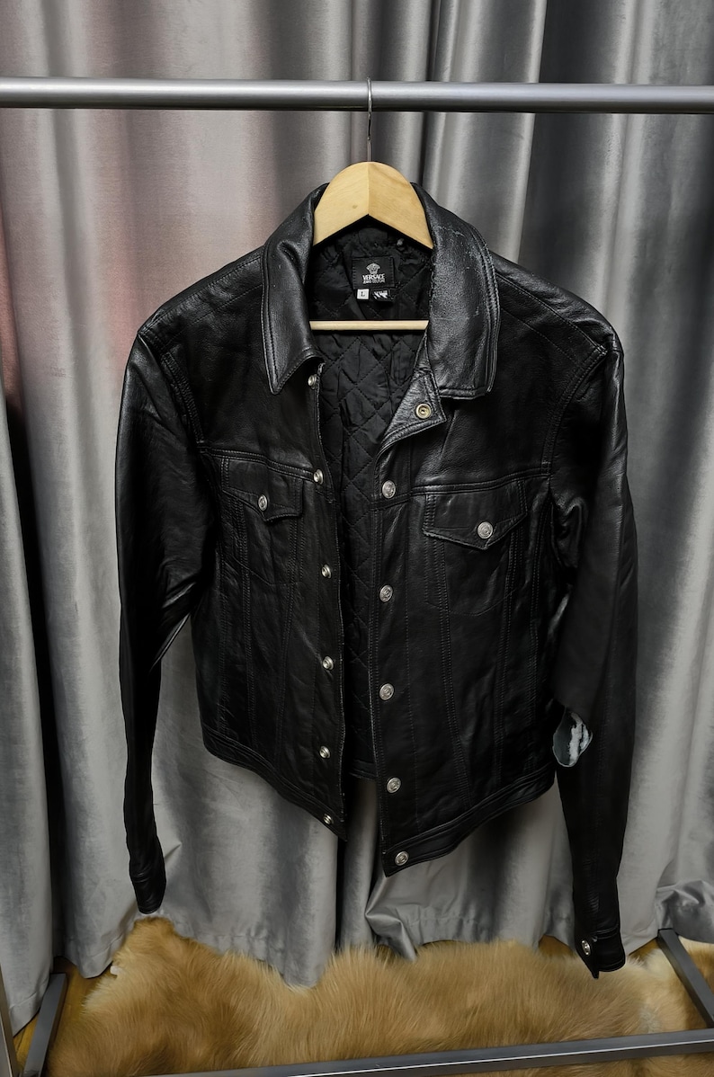 Puede incluir: Una chaqueta de cuero negra con cuello en punta, mangas largas y cierre frontal de botones de metal plateado. La chaqueta incluye dos bolsillos en el pecho con solapas abotonadas y un forro interior negro acolchado.