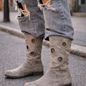 Peut inclure: Bottes en daim gris clair avec des découpes circulaires. Portées avec un jean gris délavé. Les bottes ont un petit talon et un bout arrondi. Les bottes mesurent environ 30 cm de hauteur.