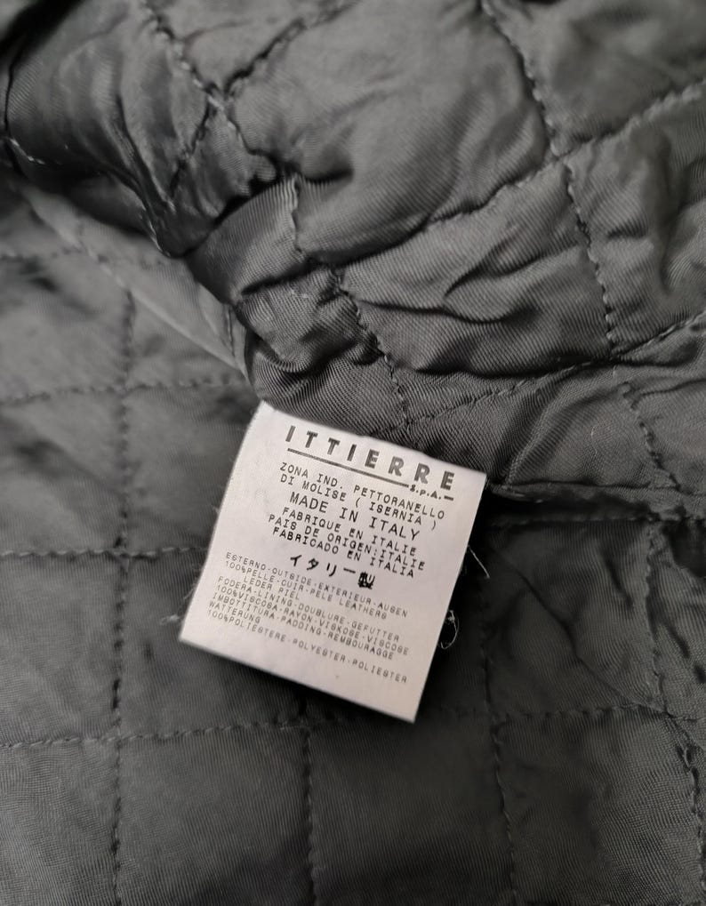 Puede incluir: Un interior de prenda acolchada negra con una etiqueta blanca rectangular de cuidado y origen. La etiqueta muestra el texto ITTIERRE S.p.A., Made in Italy, y enumera composiciones de materiales que incluyen 100% cuero exterior, 100% forro de viscosa y 100% relleno de poli&eacute;ster.