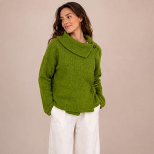 Op de afbeelding: Een groene wikkel trui met een brede kraag en een knoopsluiting. De trui heeft lange mouwen en een striksluiting aan de zijkant. Het model draagt een witte broek.