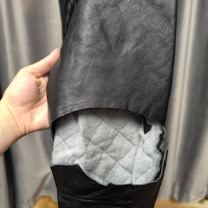 Puede incluir: Una chaqueta de cuero negro con un forro interior visible. La capa exterior es de cuero negro liso, mientras que la secci&oacute;n interior expuesta revela un forro de tela acolchada de color gris claro con un patr&oacute;n de costuras en forma de diamante.
