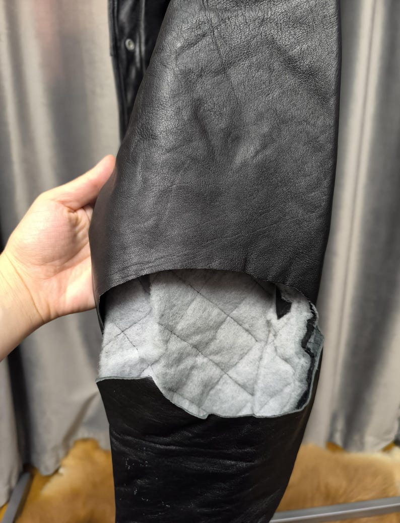 Puede incluir: Una chaqueta de cuero negro con un forro interior visible. La capa exterior es de cuero negro liso, mientras que la secci&oacute;n interior expuesta revela un forro de tela acolchada de color gris claro con un patr&oacute;n de costuras en forma de diamante.