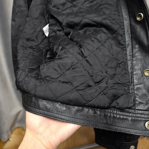 Puede incluir: Una chaqueta de cuero negro con forro interior acolchado con un patr&oacute;n de costuras en forma de diamante. La chaqueta incluye un cierre frontal con botones de presi&oacute;n met&aacute;licos a lo largo de la solapa y una etiqueta de cuidado blanca.