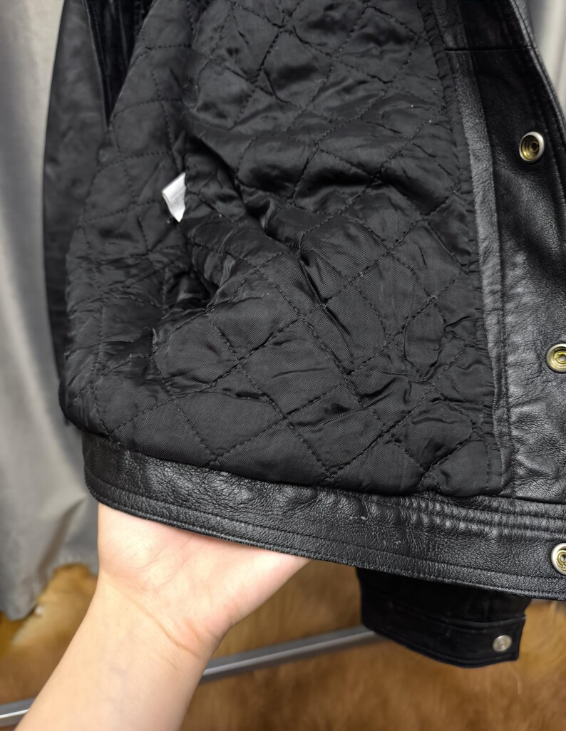 Puede incluir: Una chaqueta de cuero negro con forro interior acolchado con un patr&oacute;n de costuras en forma de diamante. La chaqueta incluye un cierre frontal con botones de presi&oacute;n met&aacute;licos a lo largo de la solapa y una etiqueta de cuidado blanca.