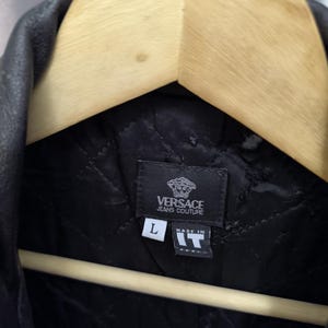 Puede incluir: Una chaqueta negra acolchada con una etiqueta de Versace Jeans Couture. La etiqueta interior muestra la letra L de talla y una etiqueta que dice Made in IT. La prenda cuelga de una percha de madera, mostrando un patr&oacute;n de costura en rombos.