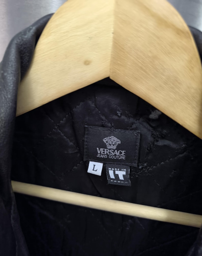 Puede incluir: Una chaqueta negra acolchada con una etiqueta de Versace Jeans Couture. La etiqueta interior muestra la letra L de talla y una etiqueta que dice Made in IT. La prenda cuelga de una percha de madera, mostrando un patr&oacute;n de costura en rombos.
