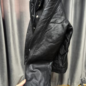 Puede incluir: Una chaqueta de cuero negro con cuello cl&aacute;sico, mangas largas con pu&ntilde;os abotonados y cierre frontal de botones. La chaqueta presenta detalles de costuras visibles a lo largo de las mangas y el torso, con un acabado liso y texturizado.