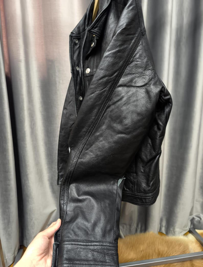 Puede incluir: Una chaqueta de cuero negro con cuello cl&aacute;sico, mangas largas con pu&ntilde;os abotonados y cierre frontal de botones. La chaqueta presenta detalles de costuras visibles a lo largo de las mangas y el torso, con un acabado liso y texturizado.