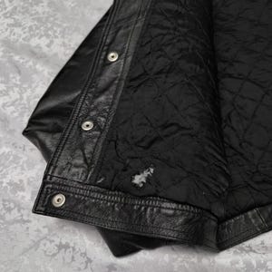 Puede incluir: Una chaqueta de cuero negro con una tapeta frontal con botones de presi&oacute;n de metal plateado. El interior est&aacute; forrado con tela acolchada negra, que muestra una peque&ntilde;a zona de desgaste visible cerca del dobladillo inferior de la prenda.