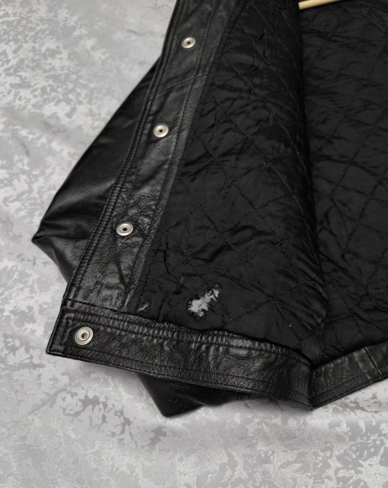 Puede incluir: Una chaqueta de cuero negro con una tapeta frontal con botones de presi&oacute;n de metal plateado. El interior est&aacute; forrado con tela acolchada negra, que muestra una peque&ntilde;a zona de desgaste visible cerca del dobladillo inferior de la prenda.