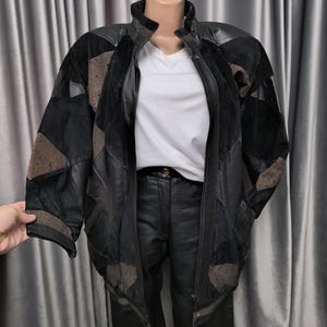 Peut inclure: Veste en cuir noir avec un design patchwork et des empiècements en daim marron. La veste a une fermeture éclair et un col montant. Elle est présentée sur un mannequin avec un t-shirt blanc et un pantalon noir.