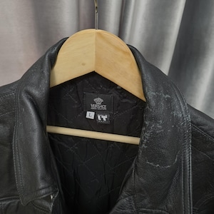Puede incluir: Una chaqueta de cuero negro con forro interior acolchado y cuello en punta. La etiqueta interior muestra el logotipo de Versace Jeans Couture, una etiqueta de talla L y una etiqueta Made in IT. El cuero muestra signos visibles de desgaste.